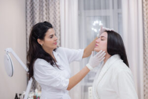 A female dermatologist in a white lab coat and gloves examines a female patient's jawline and forehead during a skin consultation in a bright, modern medical office.